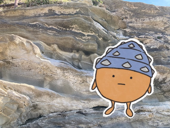 トークプログラム「ほりだせ! 地球のいま・むかし 地底人どりまるくんの探検記」のイメージ画像。急な崖を下から見上げている写真。地面とほぼ平行な地層の縞模様が見えている。画像の右側には、このトークに登場する地底人の「どりまるくん」のイラストがある。どりまるくんの体は、短い手足が生えたボールのような形で、頭にはドリルの形の大きな帽子をかぶっている。